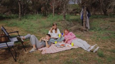 Üç çocuk, çimenli bir alanda piknik yaparken battaniyede dinlenirken, ikisi uzanmış, biri oturmuş, yiyecek ve içeceklerin keyfini çıkarırken, ağaçlar ve açık sandalyeler yakınlarda görülüyor.