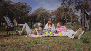 Üç genç arkadaş, ağaçlarla çevrili çimenli bir alanda piknik battaniyesine uzanmış, atıştırmalıkların ve içeceklerin keyfini çıkarıyor, neşeli ve rahat bir açık hava toplama sahnesi yaratıyorlar.