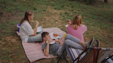 Çocuklar piknik yapmak için çimenli bir ortamda bir araya gelirler, konteynırlardaki yiyecek ve içecekleri paylaşırlar, sandalye katlayarak ve açık havada rahat bir atmosferin tadını çıkarırlar.