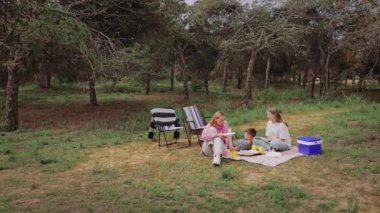 İki kadın ve bir çocuk ormanda bir battaniyenin üzerinde oturur, yiyecekleri ve kahkahaları paylaşırlar, katlanır sandalyeler ve yakınlarda yemyeşil ve arka planda ağaçlarla çevrili serinletici bir konum vardır.