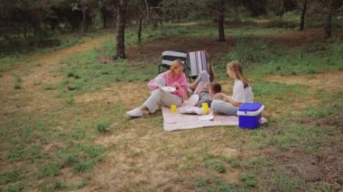 İki kadın ve bir çocuk ormanda bir piknik battaniyesinde oturup ağaçlar ve piknik malzemeleriyle çevrili yiyecek ve içecekleri paylaşıyor ve rahat bir açık hava yaratıyorlar.