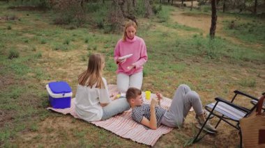 Bir grup beyaz çocuk ve kadın, çimenli bir açık alanda battaniyenin üzerinde toplandı. Soğutucudaki yiyecekleri paylaştılar. Etraflarında sandalyeler ve ağaçlar vardı. Huzurlu bir piknik sahnesi yarattılar.