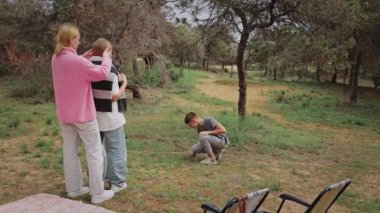 Beyaz kadın ve çocuk açık havada neşeli bir an yaşarlar, bir kadın diğerini kucaklarken çocuk yerde diz çöker ve diğer kadın kollarını neşeli bir ifadeyle kaldırır.