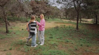 Üç genç birey, iki kadın ve bir oğlan, ağaçlar ve otlarla çevrili bir ormanda sohbet ediyorlar, çevrelerindeki doğal çevreyi ve etkileşimlerini vurguluyorlar.