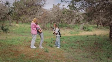 Pembe kazaklı beyaz kadın, ağaçlar ve otlarla dolu yemyeşil bir alanda başka bir çocukla uğraşırken bir çocuğu kalçasında tutuyor ve neşeli aile ilişkilerini yakalıyor.