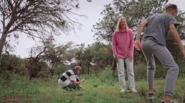 Bir grup çocuk çimenli bir alanda oyun oynarken, bir çocuk yere çömelmiş etrafı incelerken, diğer ikisi ağaçlar ve yeşillikler tarafından çerçevelenmiş eğlenceli bir anı paylaşıyor.