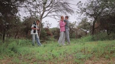Üç genç arkadaş, iki kız ve bir oğlan, yeşil bir tarlada neşeli bir an yaşarlar, bir kız diğerine sarılırken oğlan ağaçlarla ve doğal manzarayla çevrili bir şekilde ayakta durur.