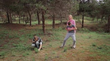 Aile bir orman ortamında açık hava keşfi yaparken, pembe kazaklı bir kadın, bir çocuğun bitkileri incelemek için çömelmesini izlerken, diğeri ağaçlar ve çimlerle çevrili olarak yakınlarda duruyor.