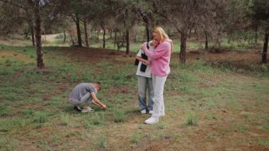 Üç birey, bir beyaz kadın ve iki çocuk yeşil bir alanda çarpışırken, bir çocuk yere odaklanmış, diğerleri ise ağaçlar ve doğal manzarayla çevrili yakın bir anı paylaşıyor.