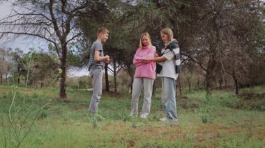 Gündelik giysiler içinde üç genç kadın bir orman açıklığında toplanıyor. Otların üzerinde dikilirken birlikte mobil bir cihazı yakından inceliyorlar. Ağaçlar doğal bir ortam sağlıyor.