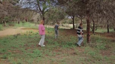 Üç kişi, pembe kazaklı bir kadın ve iki erkek çocuk, ormanlık bir alanda, ağaçlarla çevrili bitkileri ve çevreyi ve arka planda kirli bir patikayı keşfe çıkıyorlar.