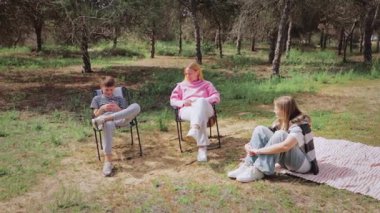 Üç genç arkadaş, açık bir ormanda güneşli bir günün tadını çıkarıyorlar. İkisi katlanır sandalyelerde, biri battaniyenin üzerinde, ağaçlarla çevrili ve yemyeşil, huzurlu bir ortamda.