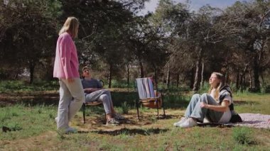 Pembe ceketli kadın, ağaçlarla çevrili çimenli bir alanda oturan iki kişiyle sohbet ediyor. Parlak güneş ışığı altında sakin bir açık hava ortamı yaratıyor.