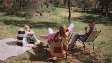 Üç genç arkadaş ormanda güneşli bir günün tadını çıkarıyorlar, sandalyeler ve battaniyeler üzerine oturmuş, doğal bir çevrede ağaçlar ve yeşilliklerle çevrili iken sohbet ve kahkahalarla meşgul oluyorlar.