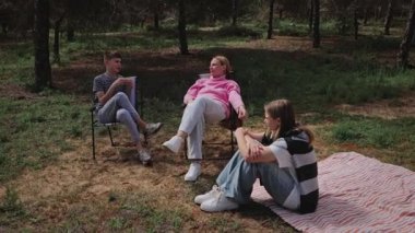 Üç genç yetişkin orman açıklığında sakin bir anın tadını çıkarırken, bir kadın sandalyede uzanırken, diğer ikisi yakınlarda, ağaçların ve desenli bir battaniyenin üzerine kurulmuş bir şekilde sohbet ediyorlar.