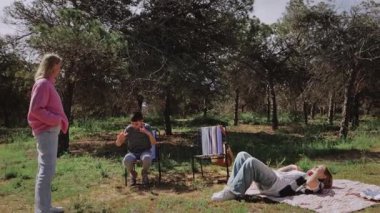 Genç arkadaşlar güneşli bir orman açıklığında sohbete dalarlar, bir kişi sandalyede oturur, diğeri battaniyeye yaslanır, ve üçüncüsü ayakta durup, gündelik ve dostane bir atmosfer yaratır.