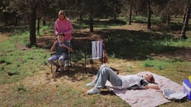 Pembe kazaklı bir kadın, oturan bir çocuğa omuz masajı yaparken, diğer bir çocuk da ağaçlarla ve yeşil otlarla dolu güneşli bir alanda battaniyede dinleniyor ve dingin bir atmosfer yaratıyor.