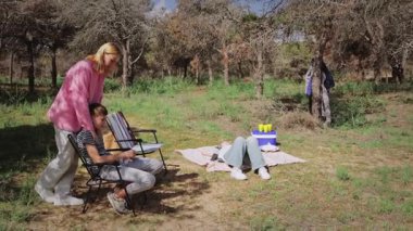 Pembe kazaklı beyaz bir kadın sandalyede oturan bir oğlanın arkasında dikilirken, bir kız çimenli bir alanda bir battaniyeye uzanmış, ağaçlar ve serin bir hava yaratıyor.