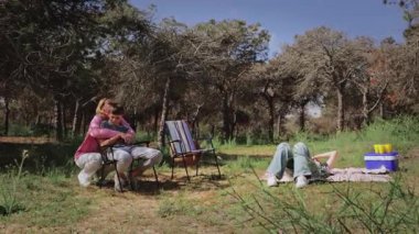 Ormanda, ağaçlar ve piknik malzemeleriyle çevrili bir şekilde battaniyeye uzanmış bir kadın sandalyeye oturmuş çocuk tutarken aile, dışarıda kaliteli zaman geçiriyor.