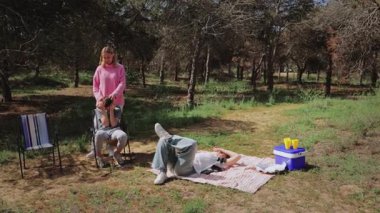 Pembe kazaklı genç beyaz kadın, bir sandalyede oturan gri kıyafetli bir kızın arkasında dikilirken, güneşli bir açık hava parkında bir oğlan, ağaçlar ve çimlerle çevrili bir şekilde bir battaniyeye yaslanıyor.