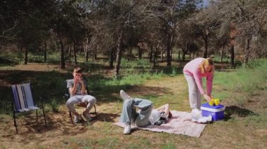 Genç yetişkinler bir orman ortamında dinlenirken, bir kişi ayakta ve elinde içki tutarken, diğer ikisi battaniyeye oturmuş, içeceklerin ve ağaçların arasından süzülen sıcak güneş ışığının tadını çıkarıyor.