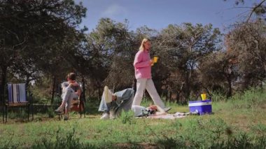 Pembe kazaklı genç beyaz kadın battaniyeye uzanmış bir arkadaşının yanında poz verirken, diğer bir arkadaşı da yeşil ormanlık alanda güneşli bir günün tadını çıkarıyor.