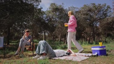 Genç yetişkinler güneşli bir günü, pembe kazaklı bir kadınla soğutucudan içecek servisi yaparak geçirirken, bir diğeri battaniyeye yaslanmış, üçüncüsü de sandalyede içkinin keyfini çıkarıyor.