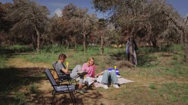 İki genç yetişkin ormanda bir battaniyenin üzerinde rahatlar, çimlerin üzerinde otururken içkilerinin tadını çıkarırlar, ağaçlar ve yeşillikler etraflarında sakin bir açık hava ortamı oluştururlar.