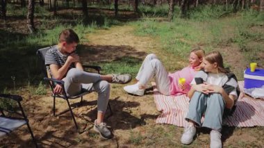 Üç genç arkadaş ormanda birlikte vakit geçirirken, bir erkek bir sandalyede oturup ayağına odaklanırken, iki kız battaniyeye uzanmış, güneşli atmosferde atıştırmalık ve içeceklerin keyfini çıkarıyorlar.