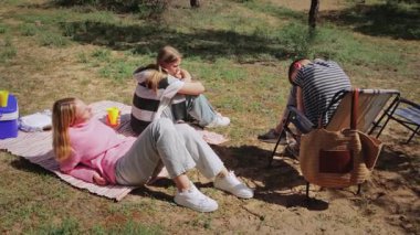 Üç genç kadın ve bir erkek çimenli bir alanda piknik battaniyesinde otururken, bir kadın içki tutuyor, bir soğutucu ve bir çanta da açık hava sahnesinin yakınına yerleştiriliyor.