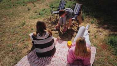 Üç genç kadın dışarıda bir battaniyenin üzerinde toplanırken, bir kadın ayakkabılarını bağlarken, diğerleri yakınlarda uzanmış, çimenli bir alanda güneşli bir günün tadını çıkarıyorlar ve katlanabilir sandalyeler mevcut.