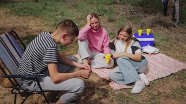 Üç genç arkadaş, iki kadın ve bir erkek, çimenli bir alanda bir battaniyenin üzerinde rahatlar, ağaçlarla çevrili, güneşli bir atmosferde sohbet eder ve içkinin tadını çıkarırlar.