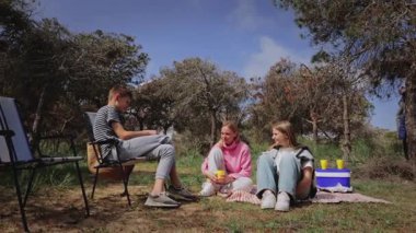 Genç arkadaşlar ormanda piknik yapmak için toplanıyorlar. İki kadın battaniyenin üzerinde ve bir adam yakınlarda oturuyor. İçkilerini paylaşıyor ve ağaçların ve yeşilliklerin arasında sohbet ediyorlar.