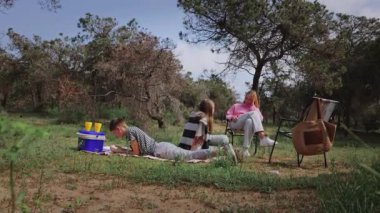 Bir grup çocuk ve bir kadın, açık havada rahatlatıcı bir günün keyfini çıkarıyorlar. Bir çocuk battaniyeye uzanmış, diğer ikisi ise sandalyeye oturmuş, ağaçlar ve çimlerle çevrili, açık bir gökyüzünün altında.