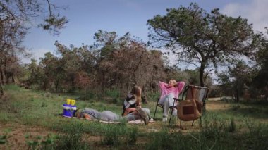 Üç kadın açık havada bir ormanda battaniyenin üzerinde dinlenirken, bir kadın yerde yatarken, diğerleri yeşil ve açık mavi gökyüzü ile çevrili sandalyelerde oturuyor.