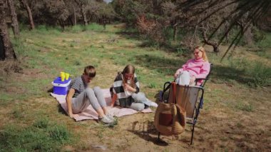 Üç genç kız güneşli bir günü ormanda, piknik malzemeleriyle bir battaniyenin üzerinde oturup, ağaçlar ve yeşilliklerle çevrili bir ortamda neşe ve neşe dolu anları paylaşarak geçiriyorlar.