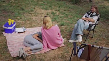 Genç kadınlar çimenli bir alanda piknik battaniyesine oturup rahatça sohbet ediyorlar. Bir kadın sandalyeye yaslanırken, diğeri de yakınlarda aperatifler ve içkilerle oturuyor.
