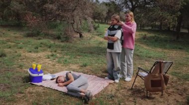 Günlük giysiler içindeki iki kadın, çimenli bir açık alanda battaniyeye uzanmış, ağaçlar ve piknik malzemeleriyle çevrili iken endişe ve etkileşim sergileyen bir arkadaşına destek oluyor.