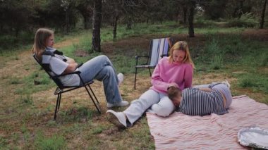 İki genç kadın rahat bir açık hava sahnesinde, bir kadın bir sandalyede ve diğeri battaniyenin üzerinde, yanında uzanan bir çocuğun kafasına şefkatle dokunuyor.