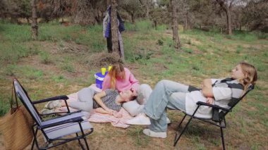 Üç genç kadın dışarıda birlikte vakit geçirirken, bir kadın battaniyenin üzerinde uzanırken, diğeri nazikçe onunla etkileşime geçiyor ve üçüncü bir kadın da ağaçlar ve çimlerle çevrili bir sandalyede oturuyor.