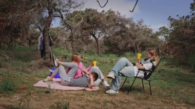 Üç genç arkadaş doğal bir ortamda battaniyeye oturmuş, içkilerini yudumluyor ve birlikte vakit geçiriyorlar, etrafları ağaçlar ve otlarla çevrili, huzurlu ve rahat bir ortam yaratıyorlar.