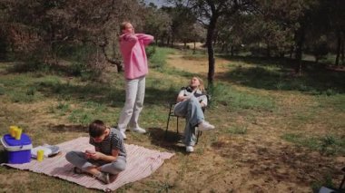Üç çocuk açık hava pikniği için bir çocuk battaniyenin üzerinde, diğeri yakınlarda, ve üçüncüsü ise ağaçlar ve piknik malzemeleriyle çevrili bir sandalyede uzanmış bir şekilde toplanıyor.