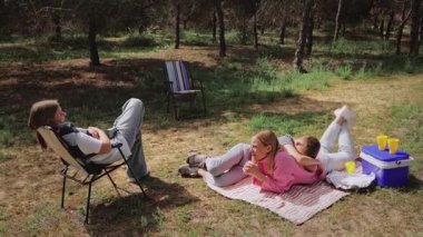 Üç genç arkadaş, güneşli bir günün tadını çıkararak ormanın ortasındaki bir battaniyeye uzanırlar. Bir kişi bir sandalyede otururken, diğer ikisi battaniyeye uzanmış soğutucuyla yakınlarda içki içiyor.