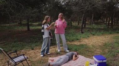 Orman ortamında battaniyenin yanında dikilen iki genç kadın, bir kadın battaniye ve sandalyelerin üzerine uzanmış ve bir soğutucu arka planda görülebiliyor.