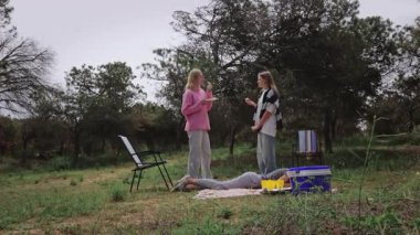İki genç kadın doğal ortamda etkileşime girer, biri yemekten zevk alırken diğeri ise piknik malzemeleri ve yeşilliklerle çevrili olarak rahat bir açık hava deneyimi yaratır.
