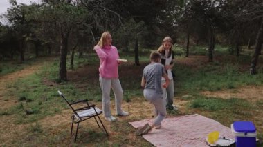 Gündelik giysiler içinde iki genç kadın açık havada canlı bir sohbete girişiyor, biri tabak tutuyor ve el kol hareketi yapıyor, genç bir adam ise yakınlarda ağaçlarla çevrili, doğal bir ortamda bir battaniyenin üzerinde oturuyor.