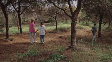 İki kadın ve bir çocuk ağaçlık alanda yürürken, bir kadın diğeriyle el ele tutuşurken, çocuk ağaçlarla çevrili ve arka planda yemyeşil çimenler arasında yürüyor.