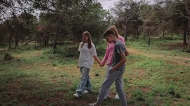 Üç birey, iki kadın ve bir erkek, ağaçlar ve yeşilliklerle dolu canlı bir ormanda el ele yürüyüp doğal ortamda barışçıl bir atmosfer yaratıyorlar.