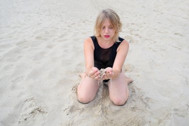 young woman kneeling in a black swimsuit holding sand in her hands