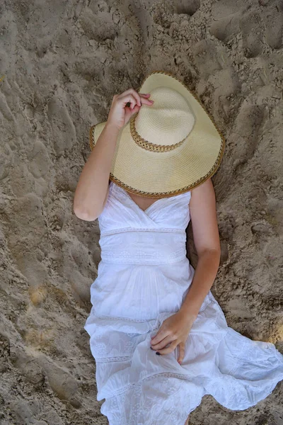 A young woman in a white dress covers her face with a hat lying on the san